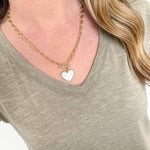 Person wearing a gold necklace with a heart pendant on a plain background