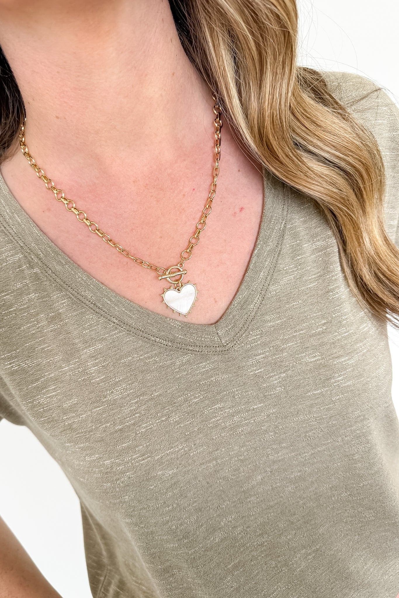 Person wearing a gold necklace with a heart pendant on a plain background