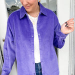 Woman wearing a purple corduroy shirt with a white top and blue jeans, standing indoors.