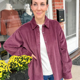 Woman wearing a purple corduroy shirt standing outside a store with flowers and a brick building in the background.