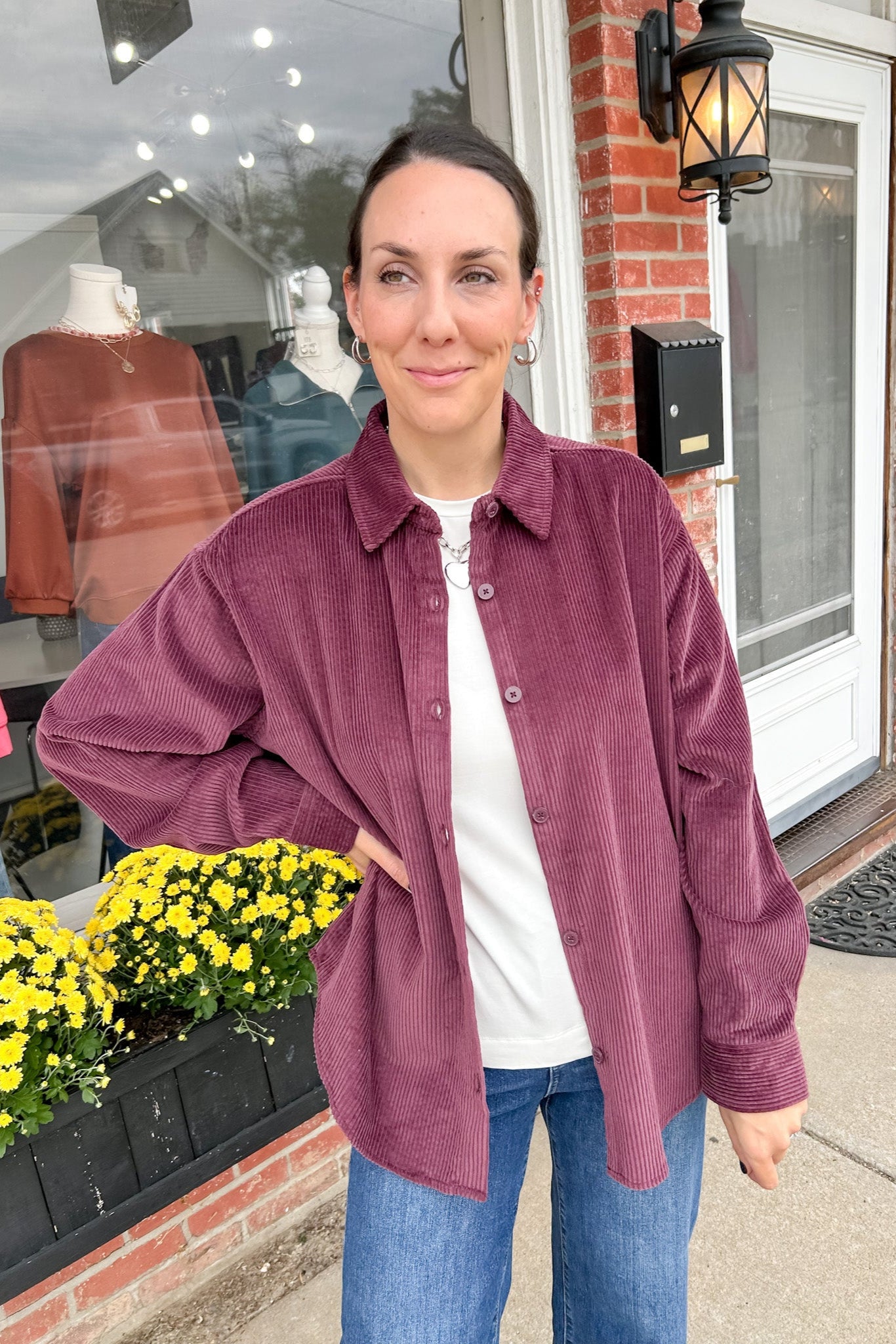 Woman wearing a purple corduroy shirt standing outside a store with flowers and a brick building in the background.