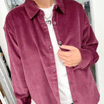 Person wearing a burgundy corduroy shirt over a white top and blue jeans indoors.