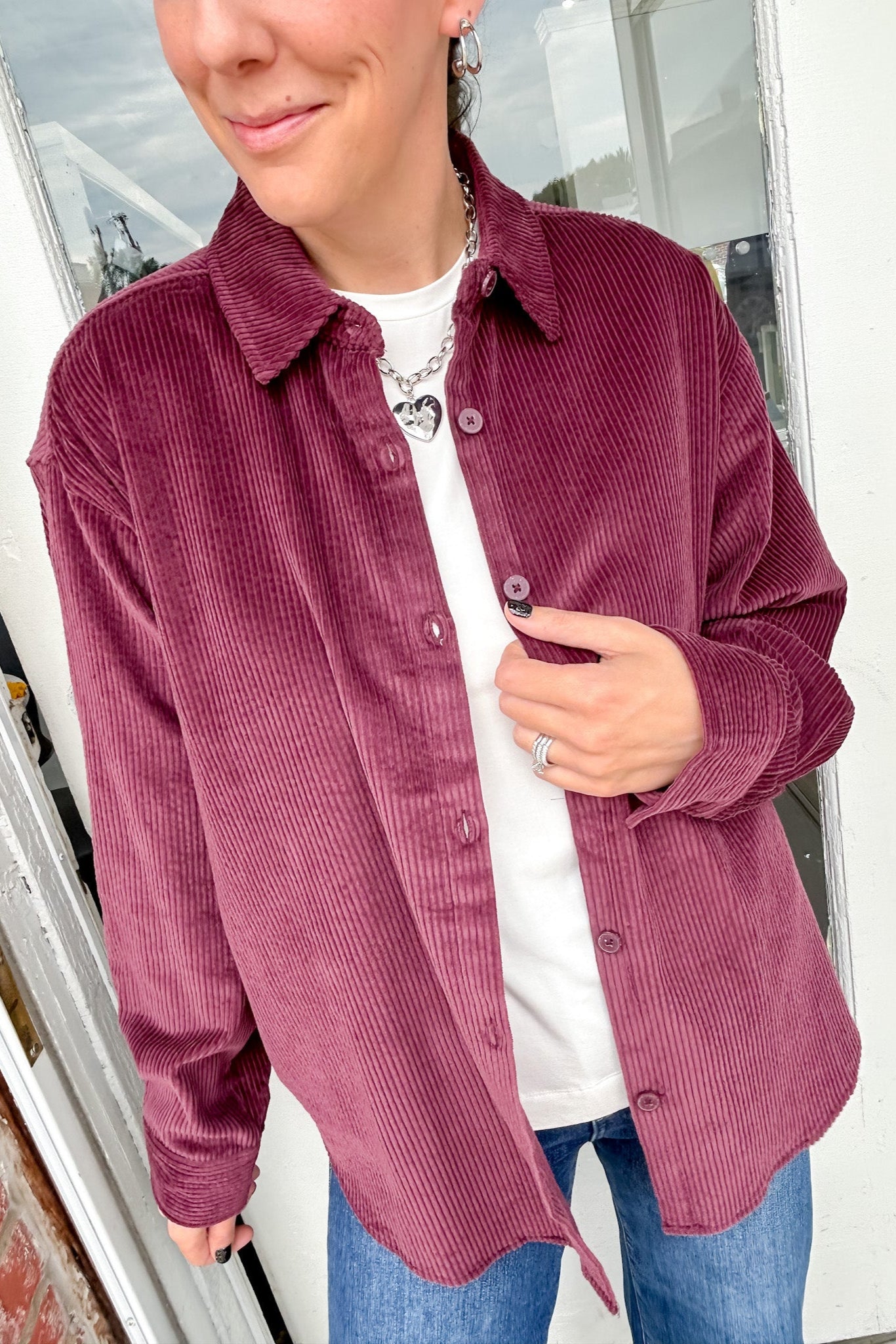 Person wearing a burgundy corduroy shirt over a white top and blue jeans indoors.