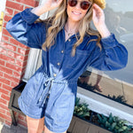 Woman wearing a blue romper and straw hat standing in front of a brick building.