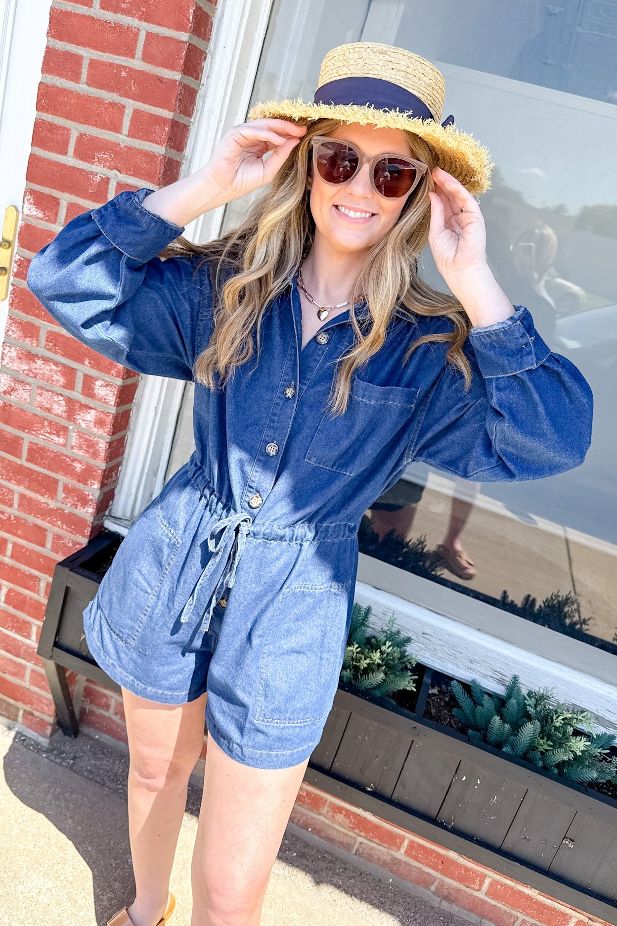 Woman wearing a blue romper and straw hat standing in front of a brick building.