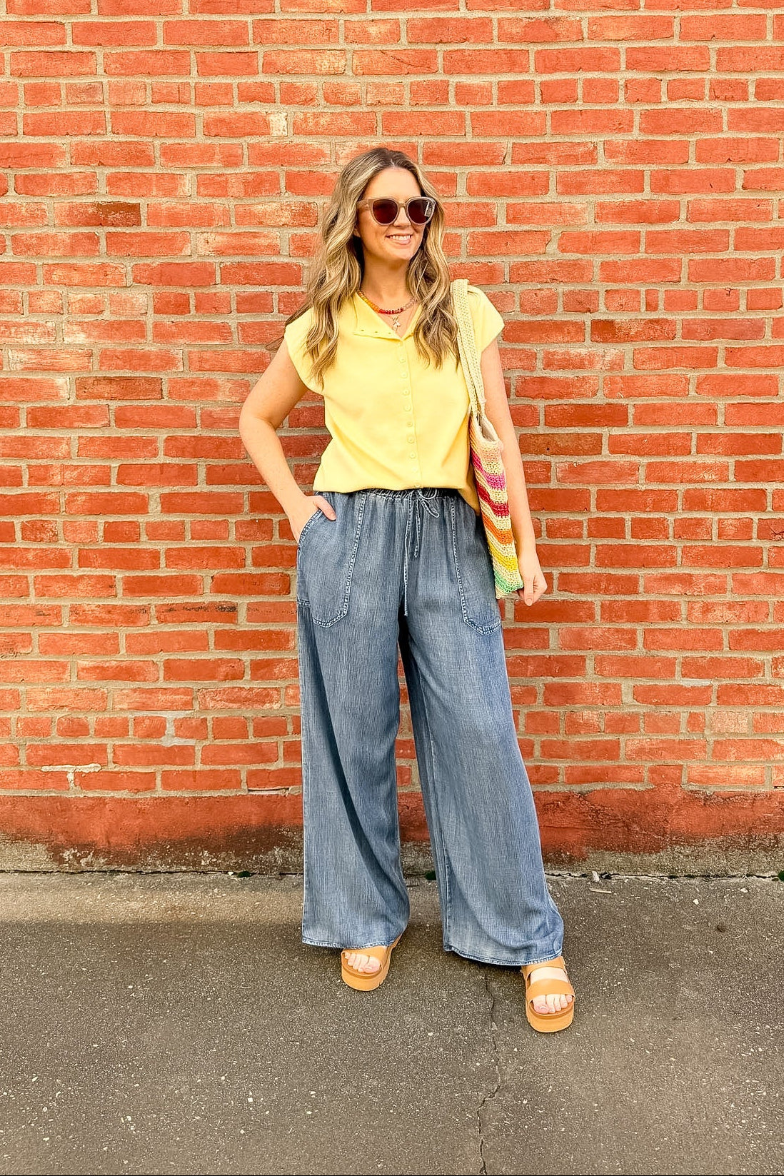Woman in yellow top and blue jeans standing against a red brick wall.