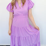 Maxi length dress with v neckline. Short sleeve with slightly bubbled sleeves. Tiered bottom. Elastic waist. Purple in color.