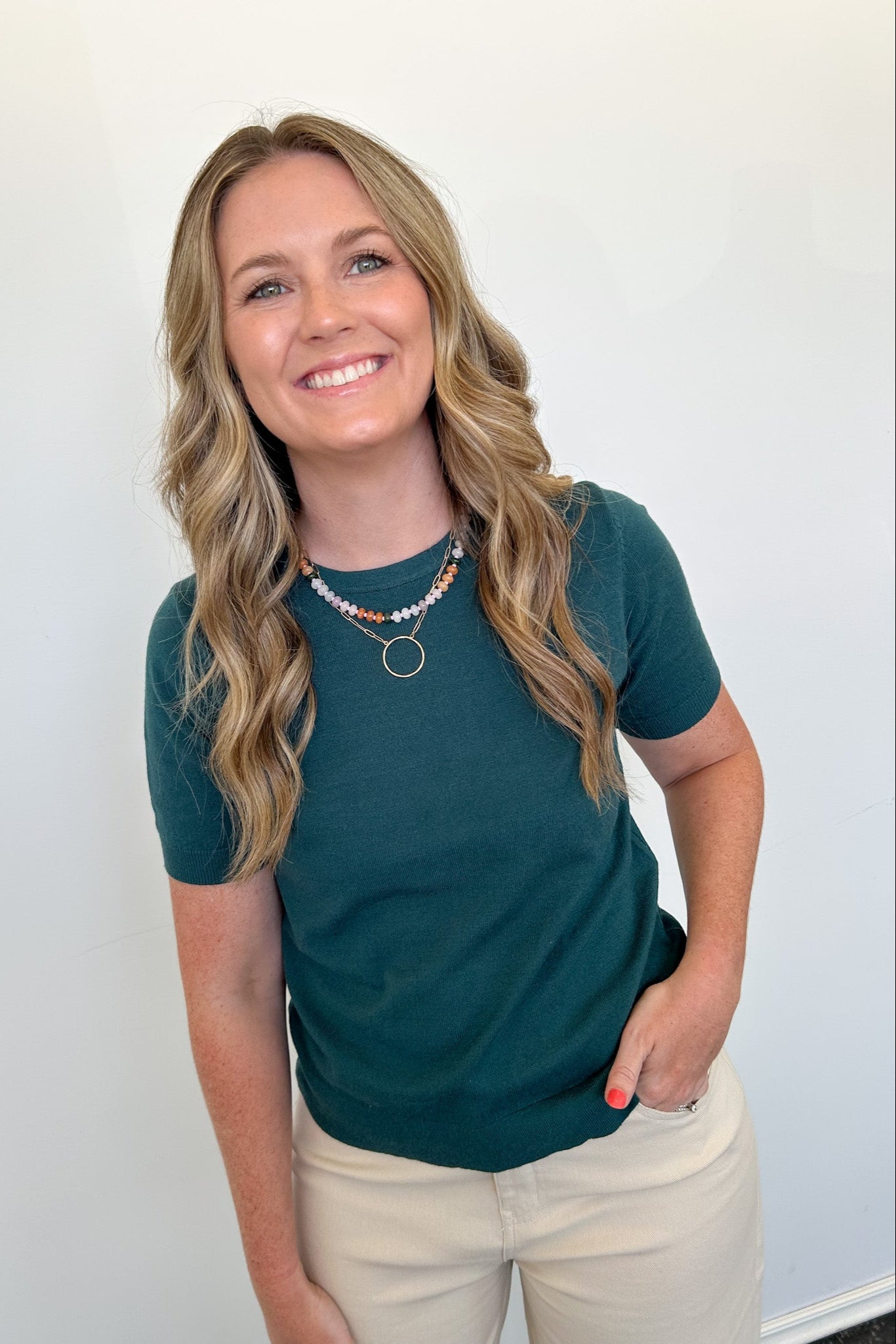 Woman wearing a teal shirt and beige pants against a white wall