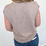 Person wearing a sleeveless brown sweater and blue jeans against a white wall.