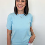 Woman wearing a light blue sweater and white pants against a plain background