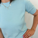 Woman wearing a light blue sweater and white pants against a plain background