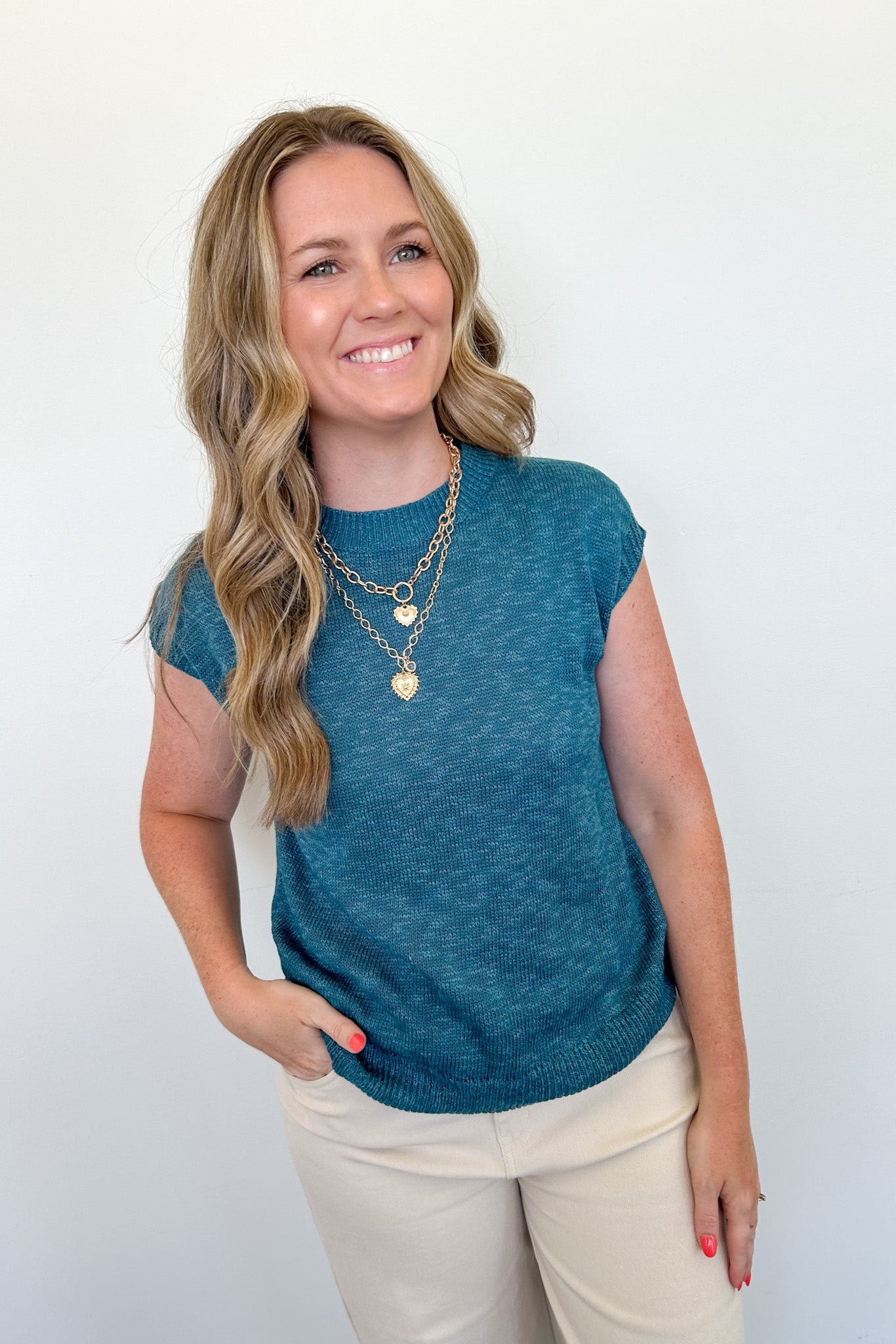 Woman wearing a teal sleeveless top with beige pants against a white background