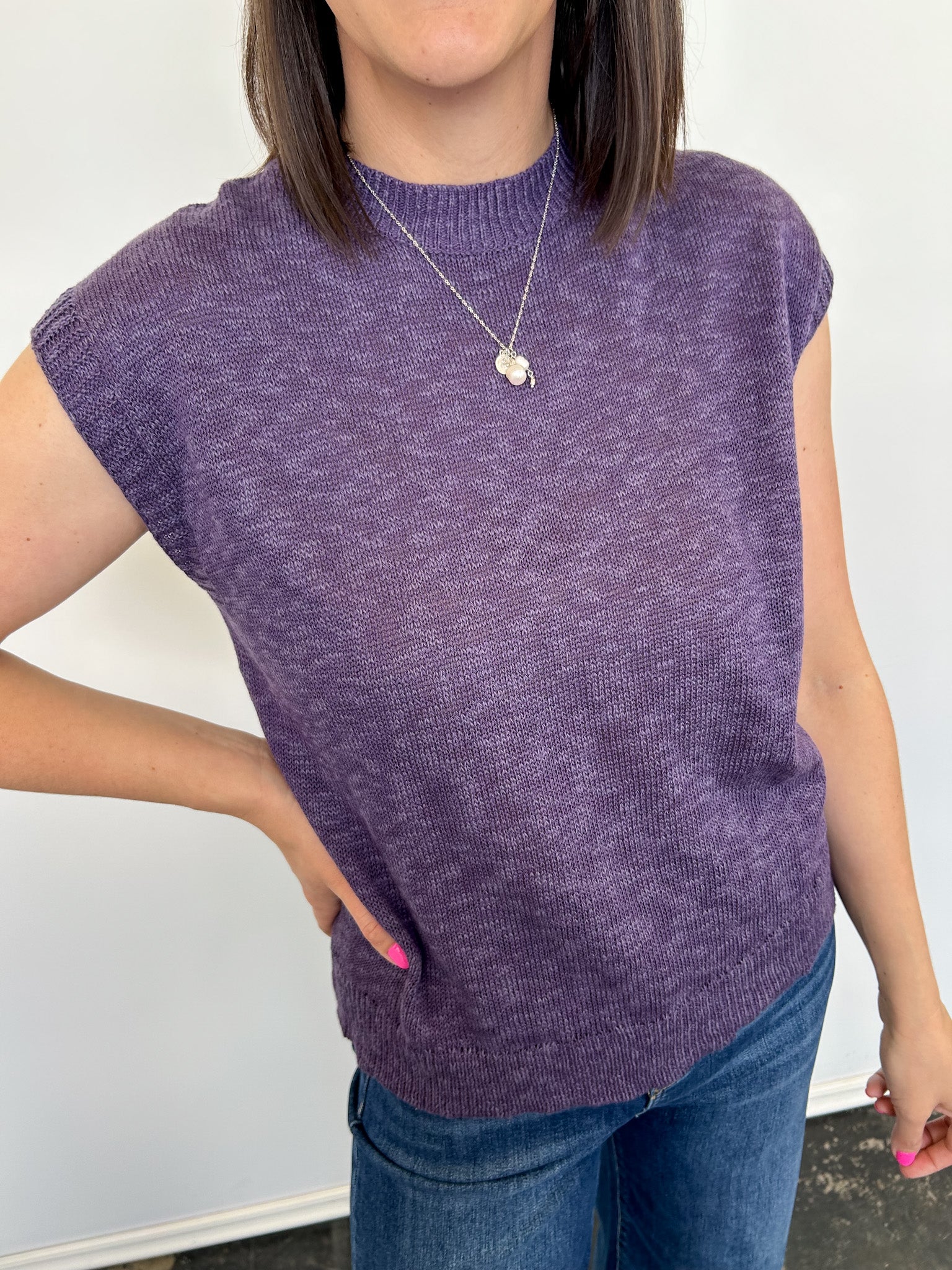 Person wearing a purple sleeveless sweater and blue jeans against a white background