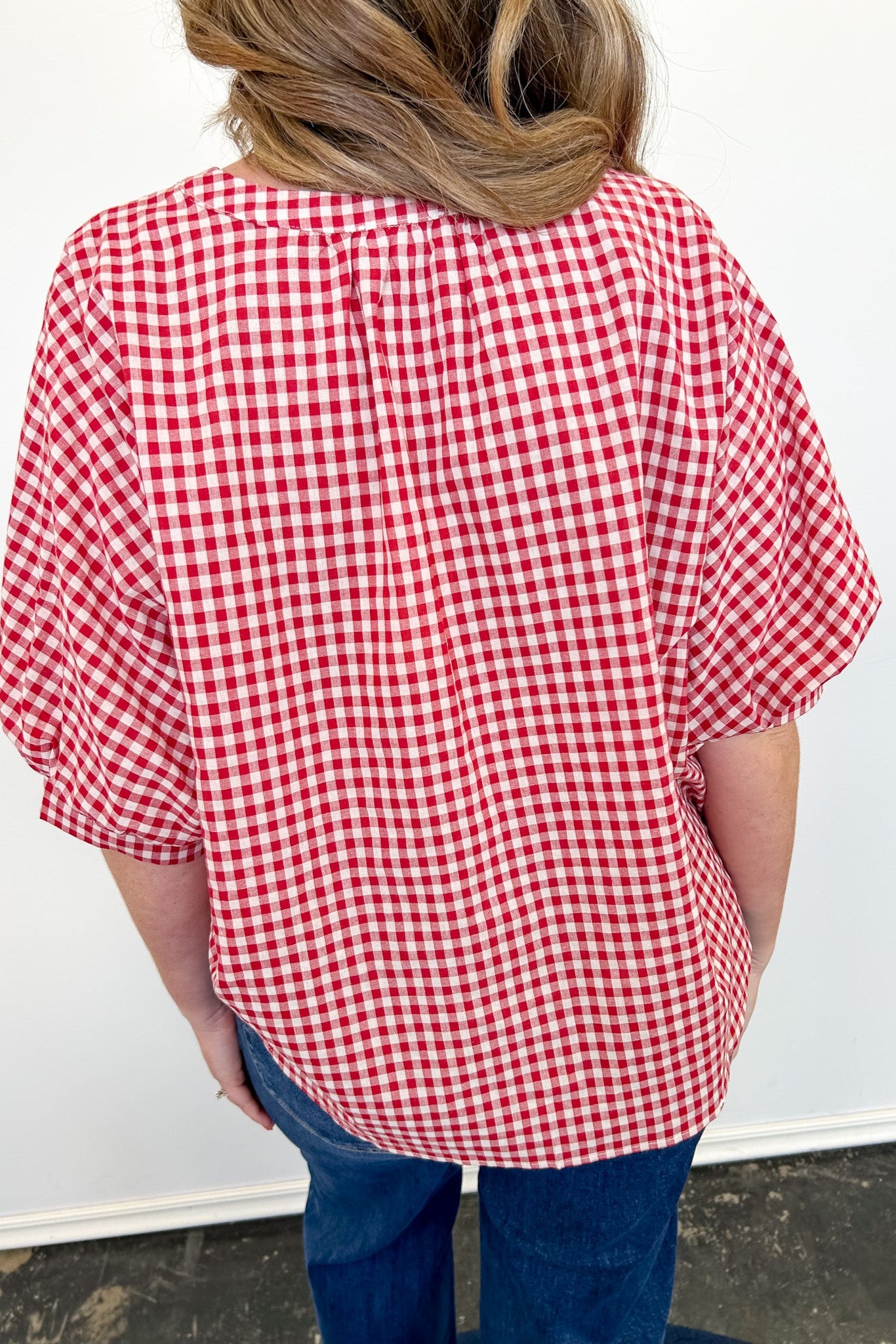 Person wearing a red and white checkered shirt with blue jeans against a white wall.