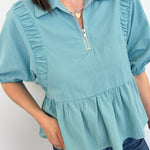 Person wearing a light blue blouse with ruffled details on a plain background