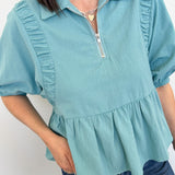 Person wearing a light blue blouse with ruffled details on a plain background