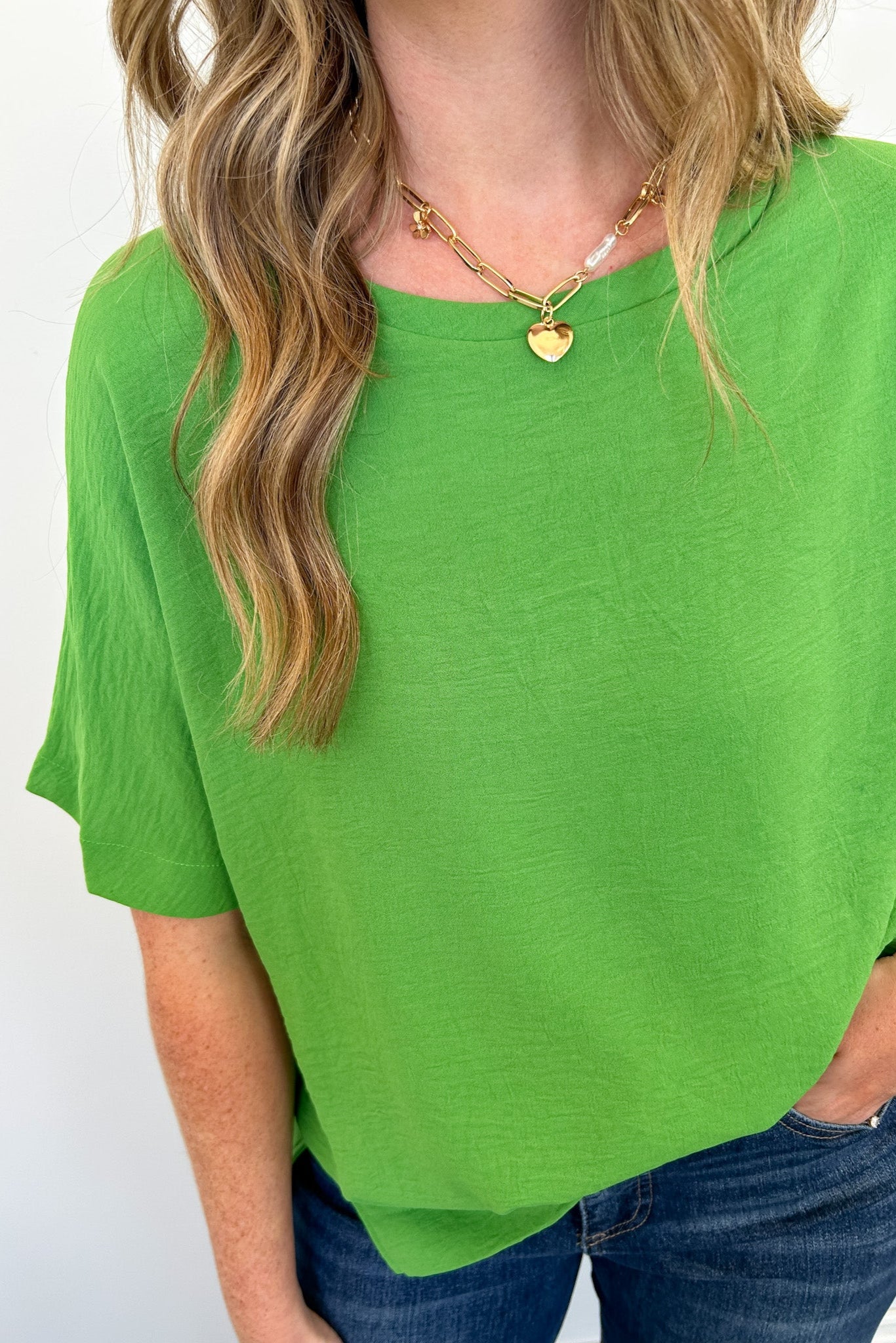 Person wearing a green top and blue jeans with a gold necklace against a white background