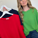 Woman holding a green top with hangers displaying red, green, and navy tops against a white background.