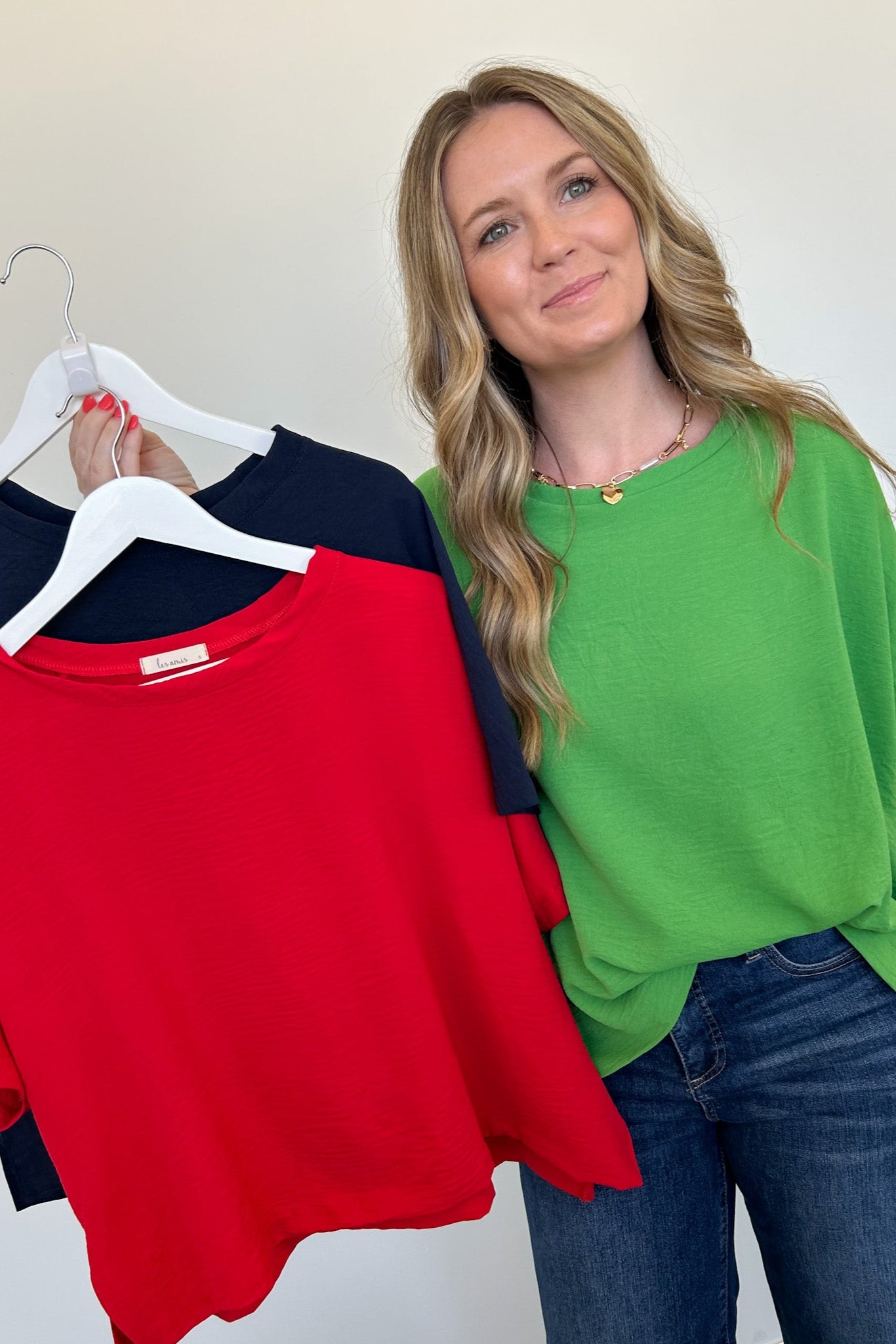 Woman holding a green top with hangers displaying red, green, and navy tops against a white background.