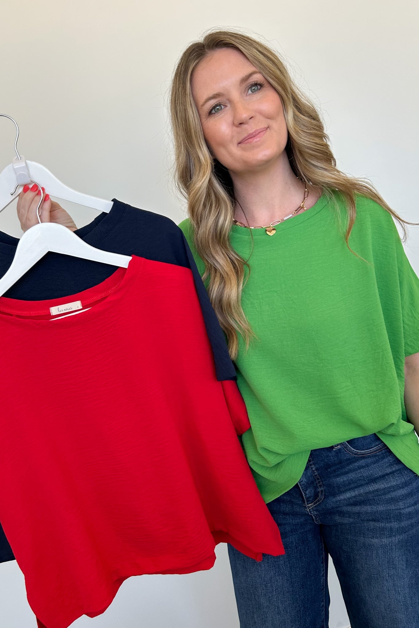 Woman holding a green top with hangers displaying red, green, and navy tops against a white background.