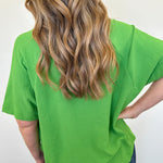 Person wearing a green t-shirt with long blonde hair, standing against a white wall.