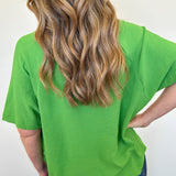 Person wearing a green t-shirt with long blonde hair, standing against a white wall.