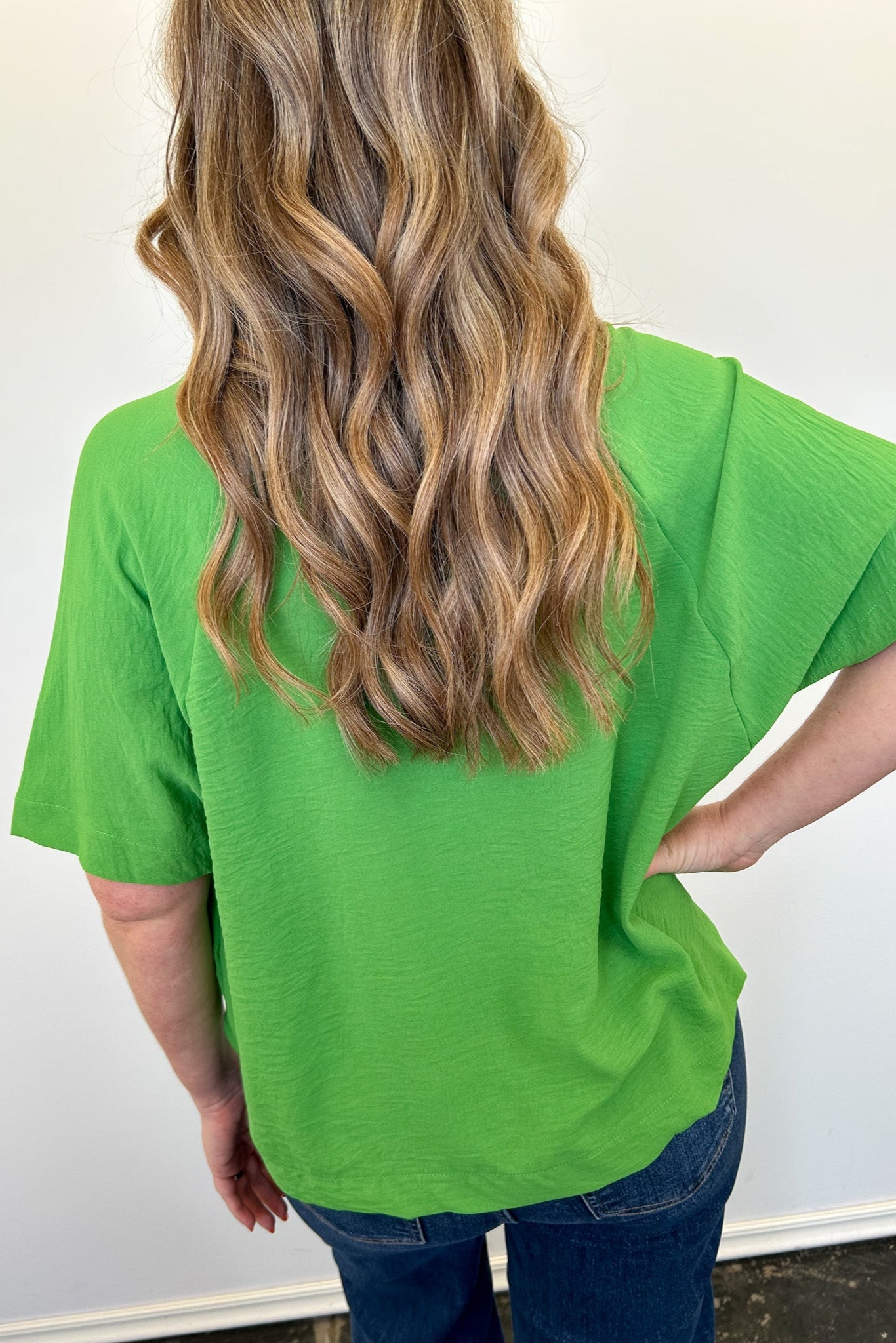 Person wearing a green t-shirt with long blonde hair, standing against a white wall.