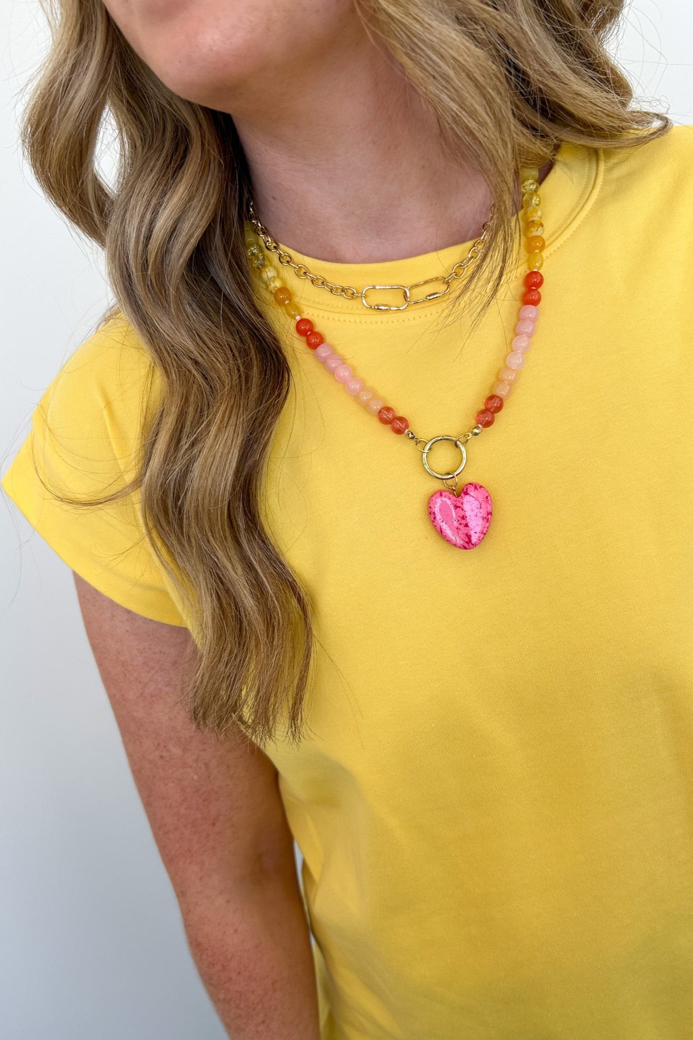 Person wearing a yellow shirt and colorful beaded necklace with a pink heart pendant on a white background