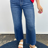 Person wearing blue jeans and white sandals on a dark blue rug with a light gray wall background