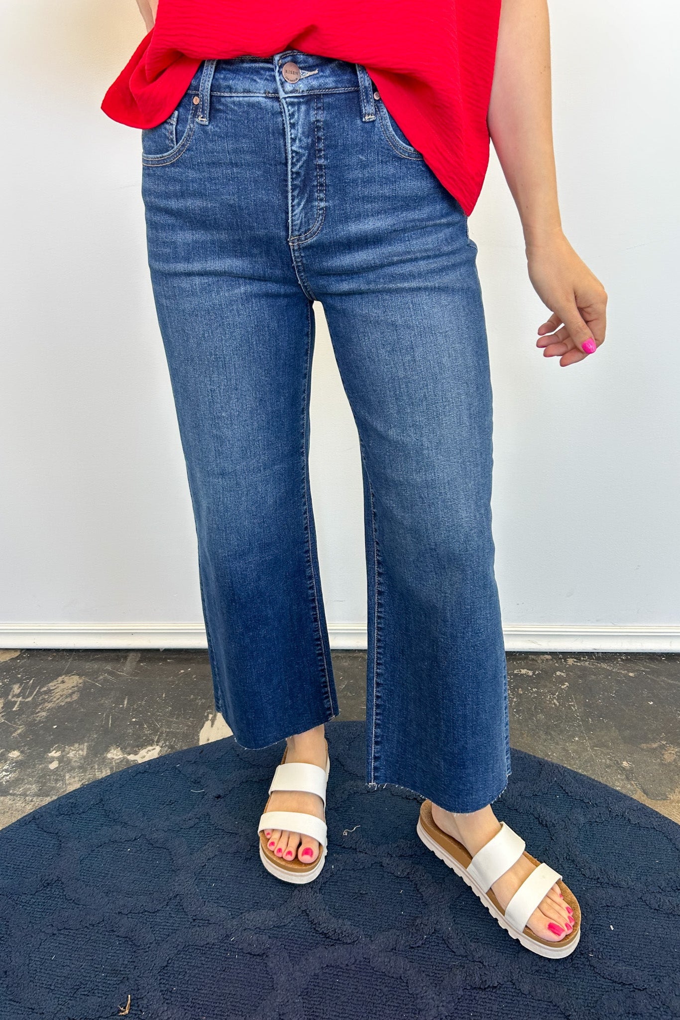 Person wearing blue jeans and white sandals on a dark blue rug with a light gray wall background