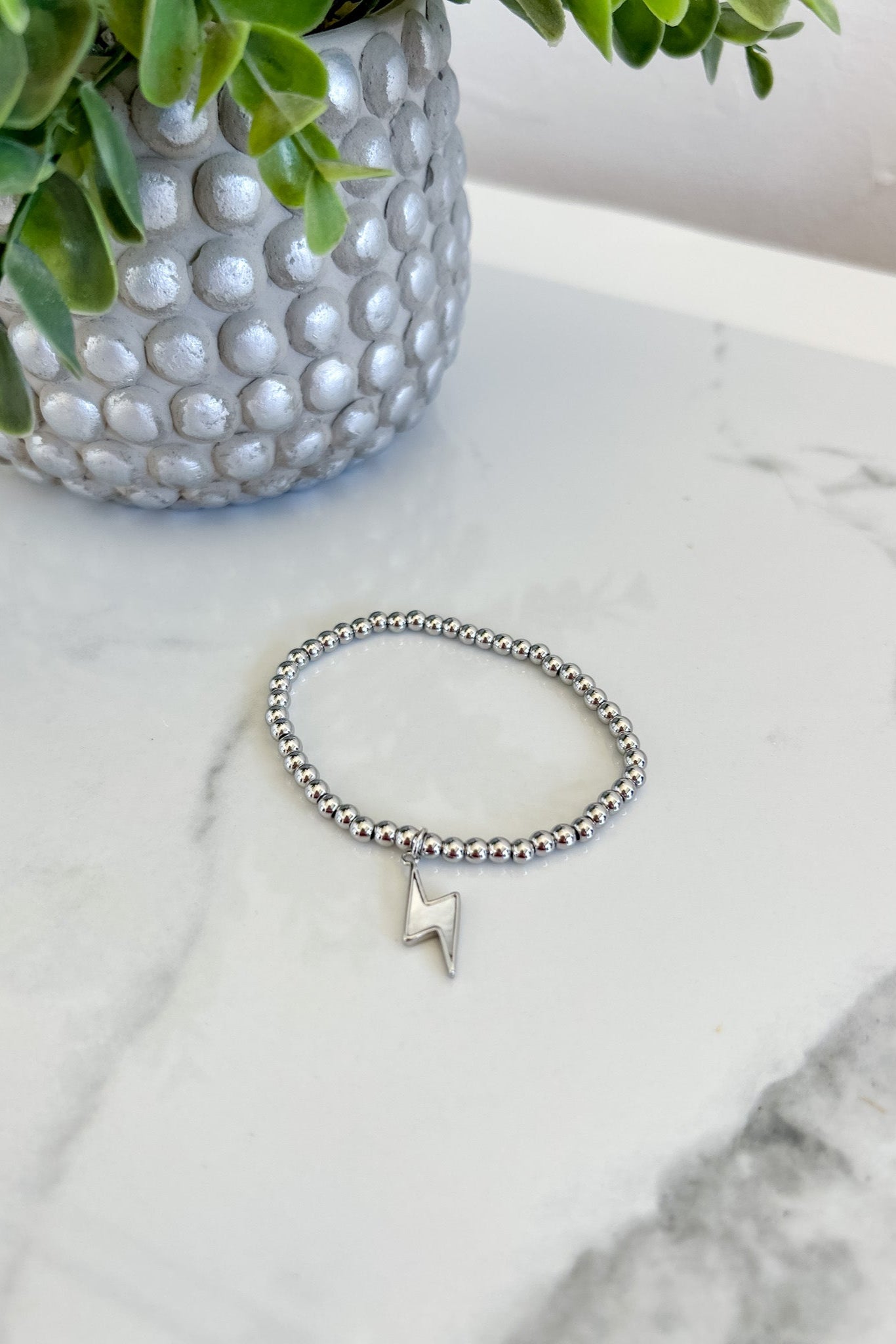 Silver bracelet with a lightning bolt charm on a marble surface next to a potted plant.