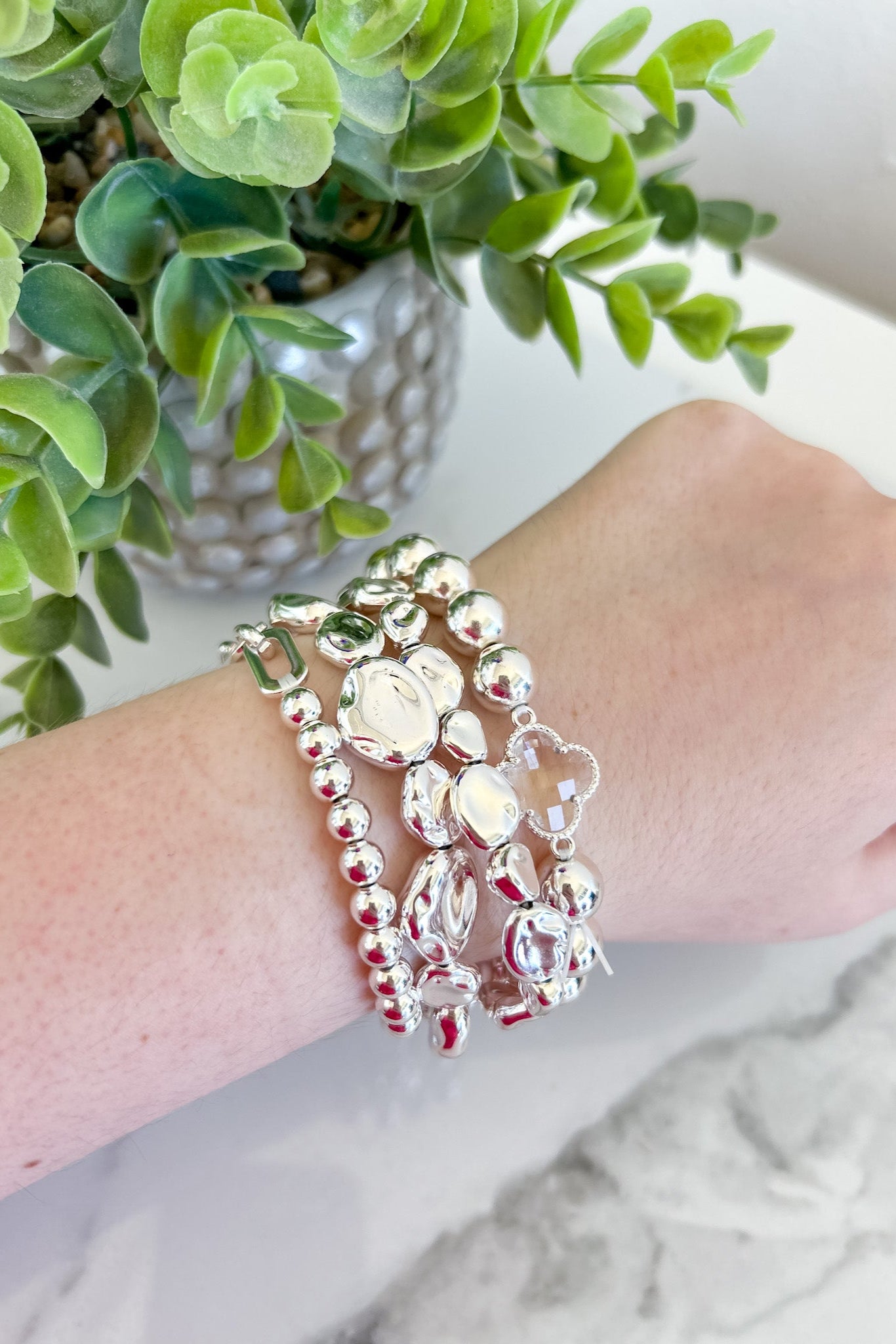 Wrist with multiple beaded bracelets against a neutral background with a plant.