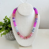 Necklace with multicolored beads on a white stand with a plant in the background