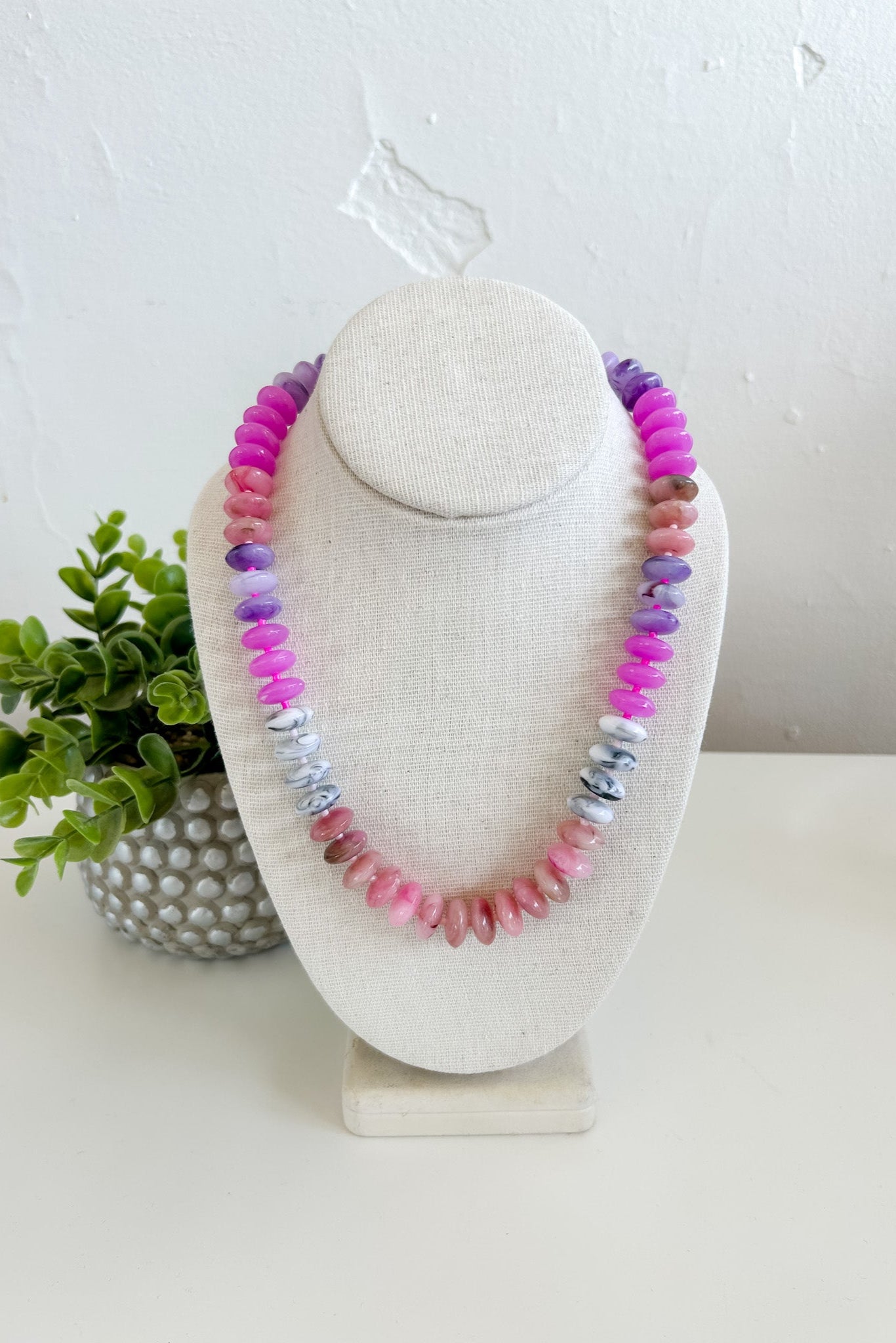 Necklace with multicolored beads on a white stand with a plant in the background