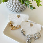 White leather earring holder with gold and silver earrings, next to a potted plant on a light surface.