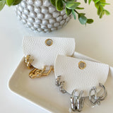 White leather earring holder with gold and silver earrings, next to a potted plant on a light surface.