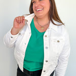 Woman wearing a white denim jacket over a green top against a plain background