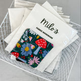 Fabric bookmark with floral design and 'milo's' branding in a wire basket.
