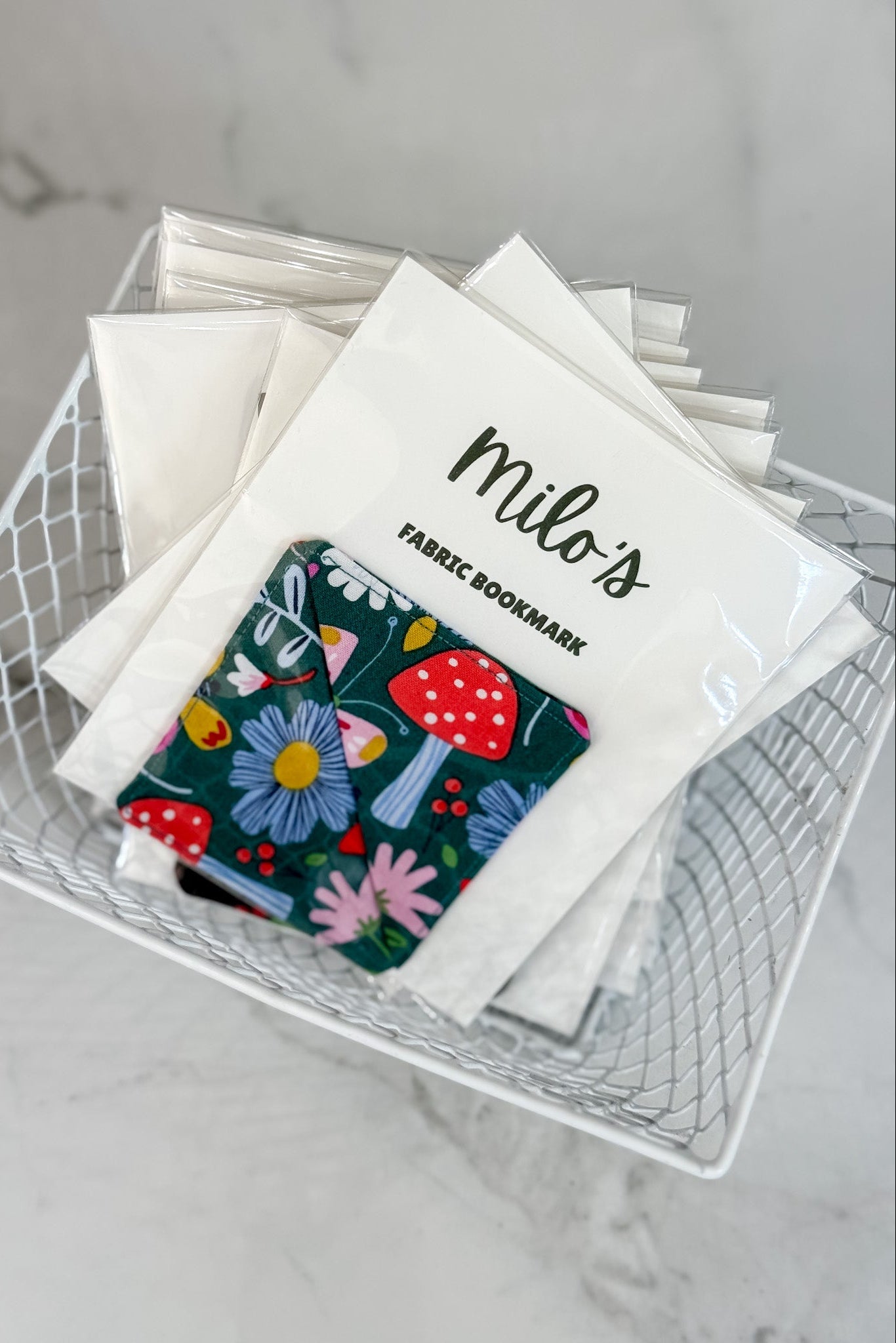 Fabric bookmark with floral design and 'milo's' branding in a wire basket.