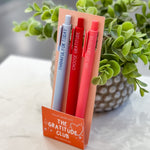 Set of pens in a holder labeled 'The Gratitude Club' on a marble surface with a plant in the background.