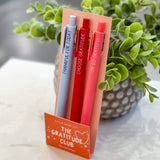 Set of pens in a holder labeled 'The Gratitude Club' on a marble surface with a plant in the background.