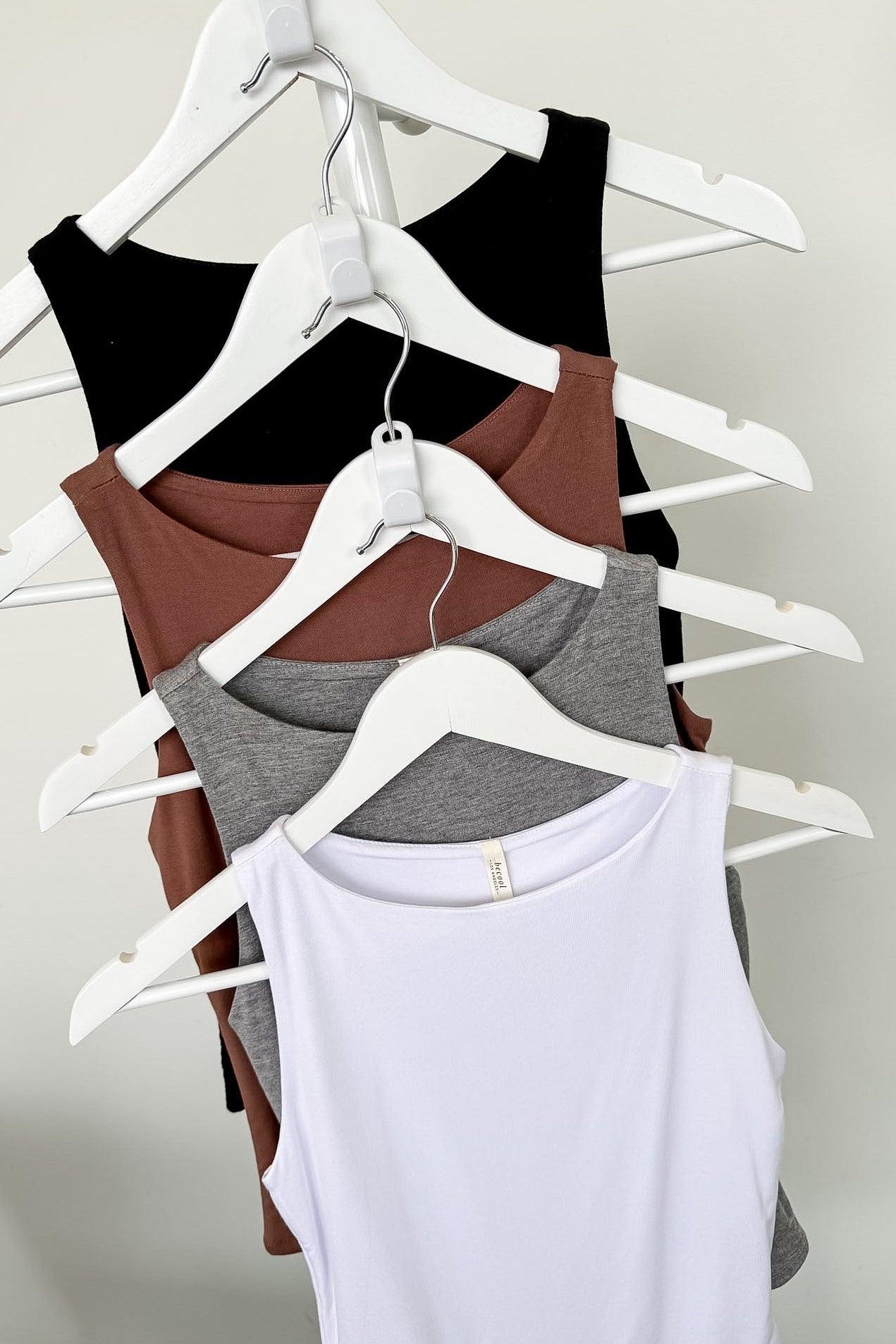 Four tank tops in black, brown, gray, and white on hangers against a plain background