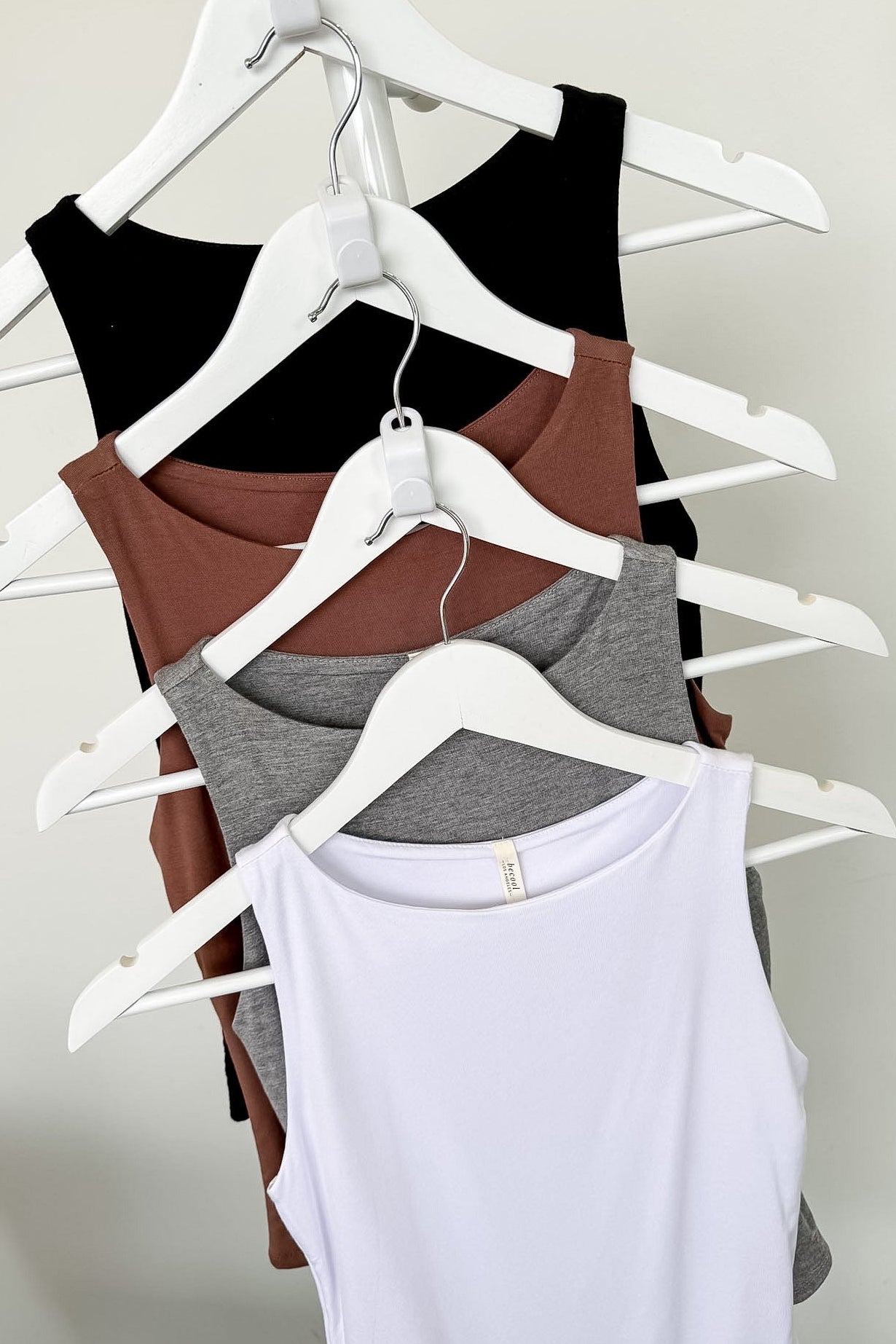 Four tank tops in black, brown, gray, and white on hangers against a plain background