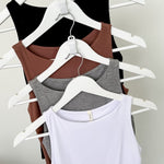 Four tank tops in black, brown, gray, and white on hangers against a plain background