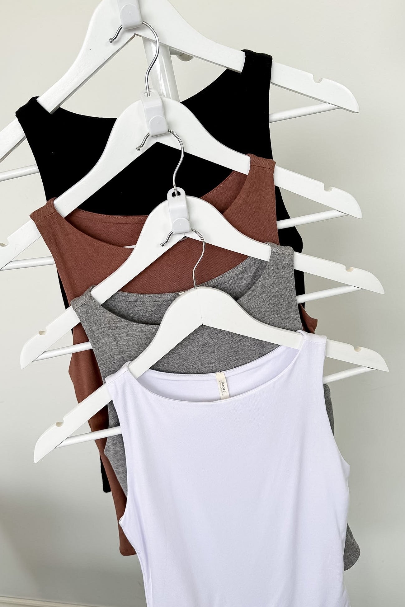 Four tank tops in black, brown, gray, and white on hangers against a plain background