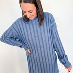 Woman wearing a short blue sweater dress against a white wall.