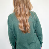 Person wearing a green sweater with long hair, standing against a white wall.