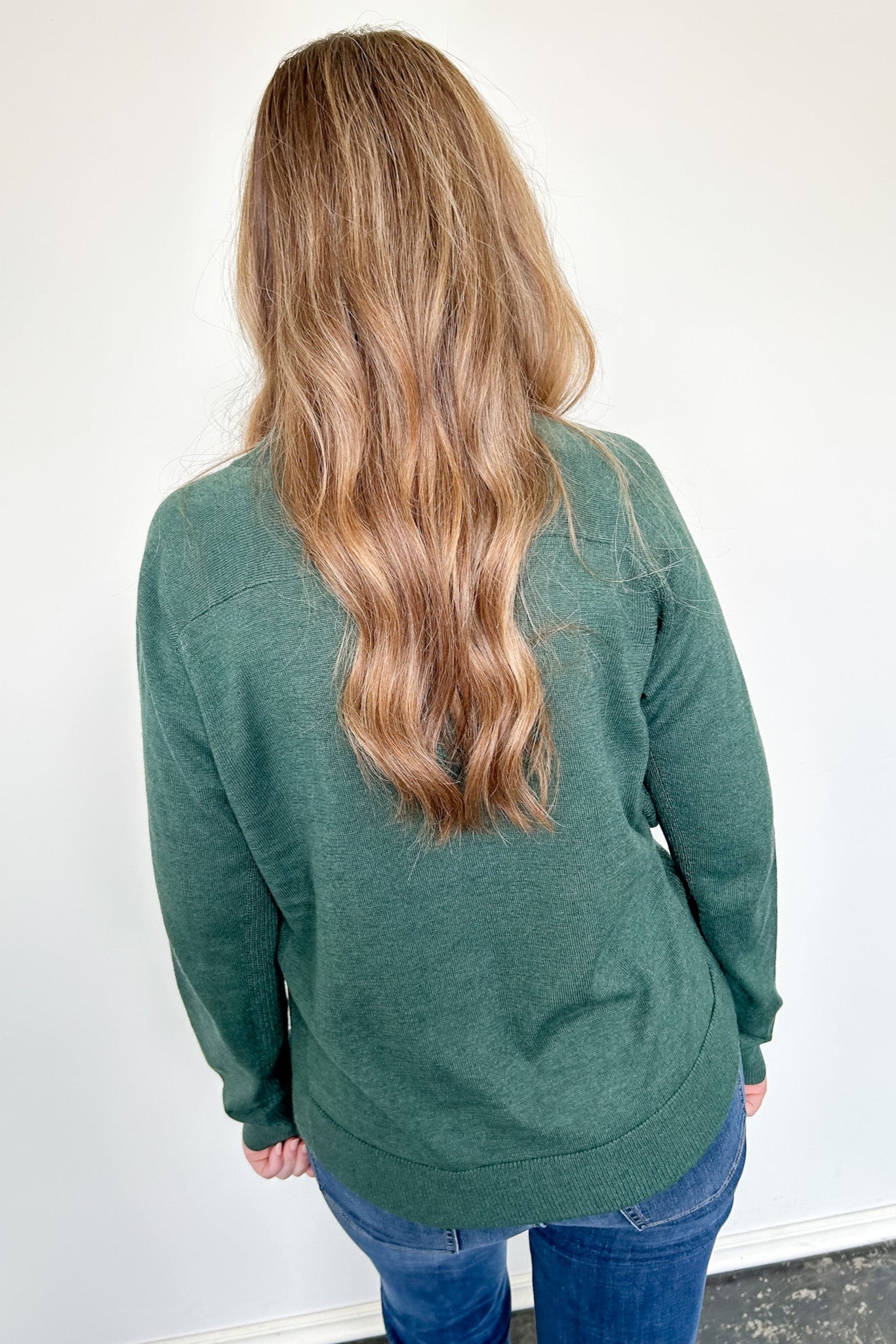 Person wearing a green sweater with long hair, standing against a white wall.