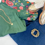 Green floral shirt, blue jeans, white sneakers, gold chain, and bracelet on a white surface.