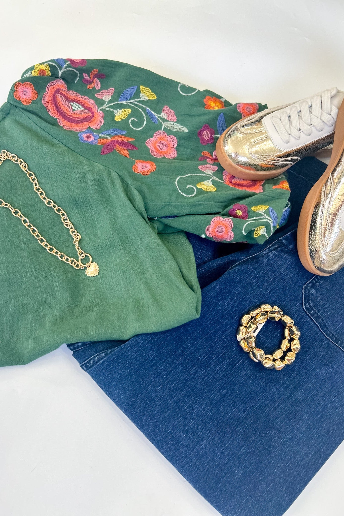 Green floral shirt, blue jeans, white sneakers, gold chain, and bracelet on a white surface.